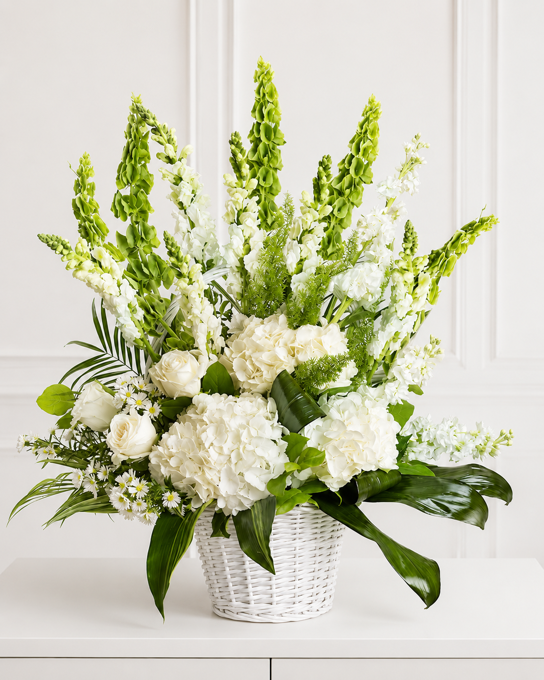 Elegante arreglo floral blanco con hortensias, rosas y flores verticales verdes en cesta blanca sobre mesa minimalista blanca.