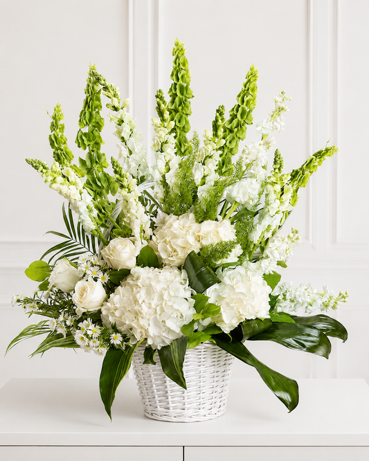 Elegante arreglo floral blanco con hortensias, rosas y flores verticales verdes en cesta blanca sobre mesa minimalista blanca.
